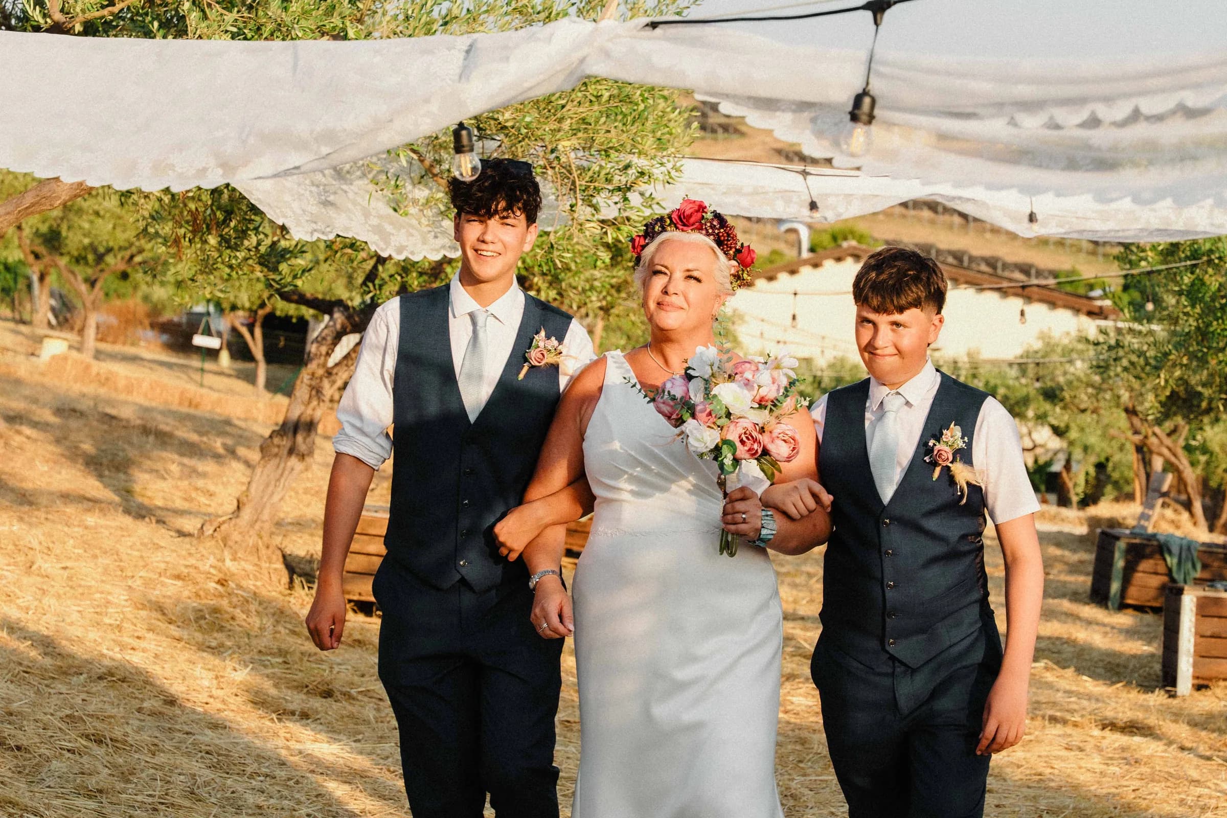 The bride walking down an aisle of dry grass and olive trees, family beside her