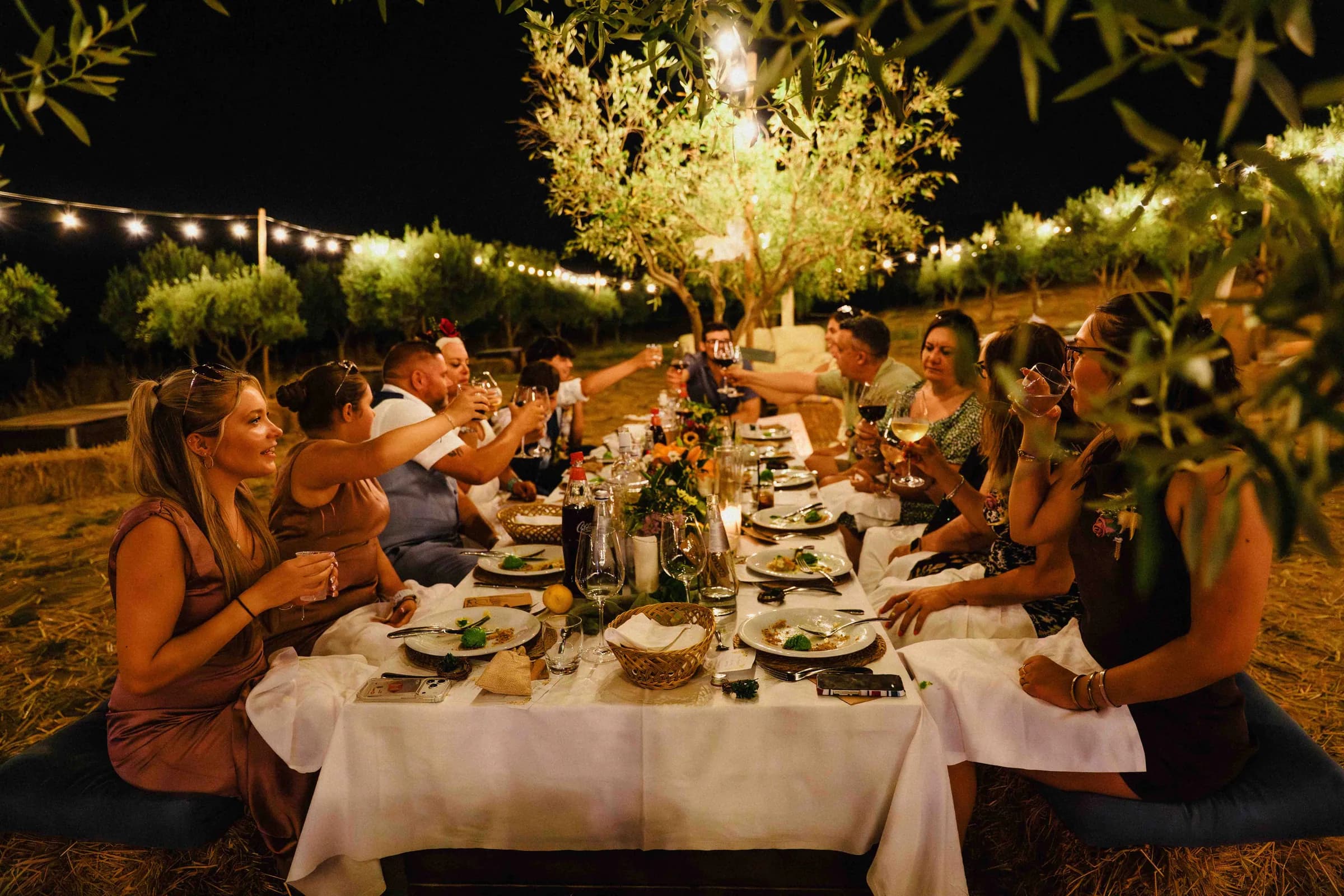 Guests at the long table after dark, warm light strung through the trees