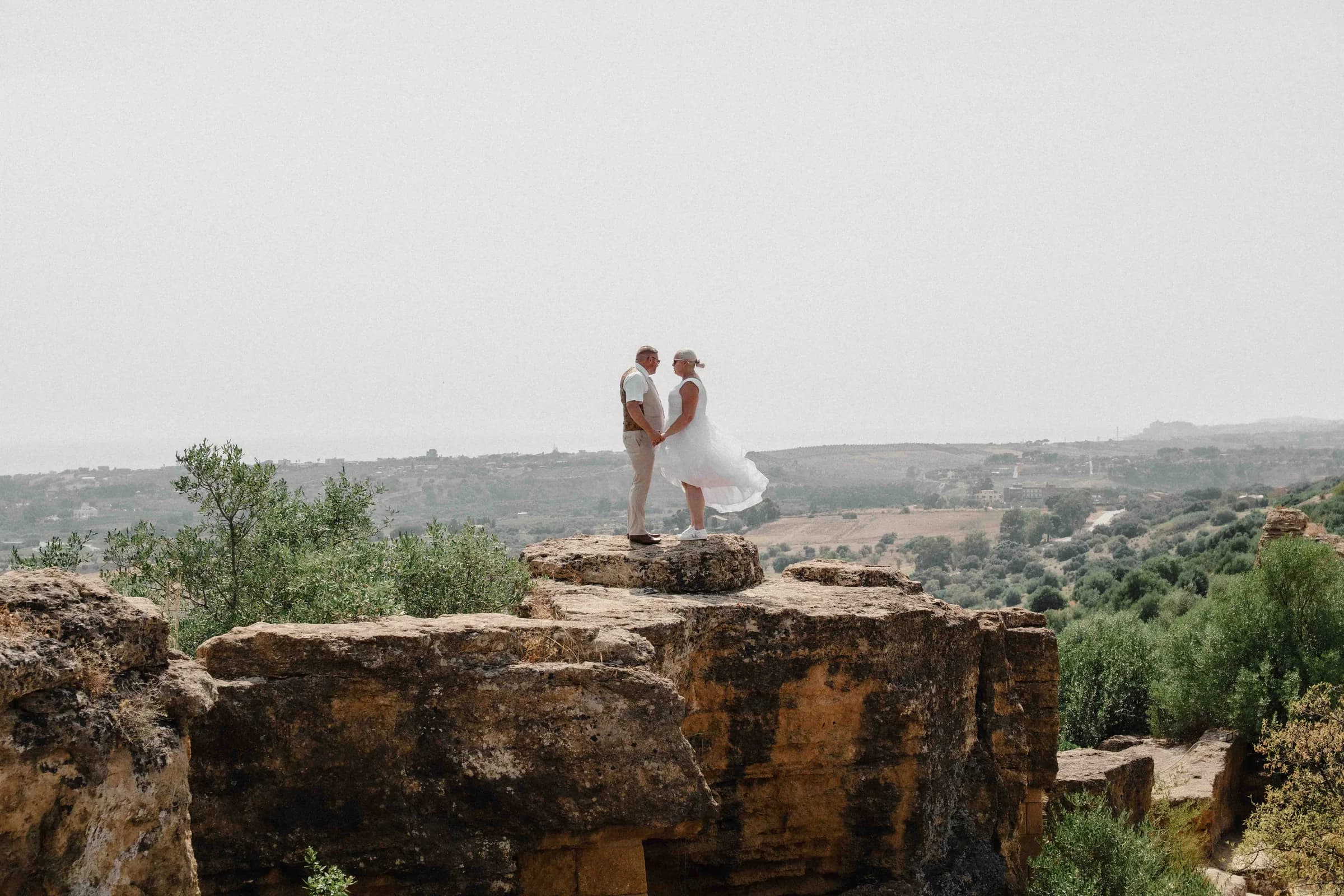 A bride and groom embracing on a weathered rock ledge, the countryside open below