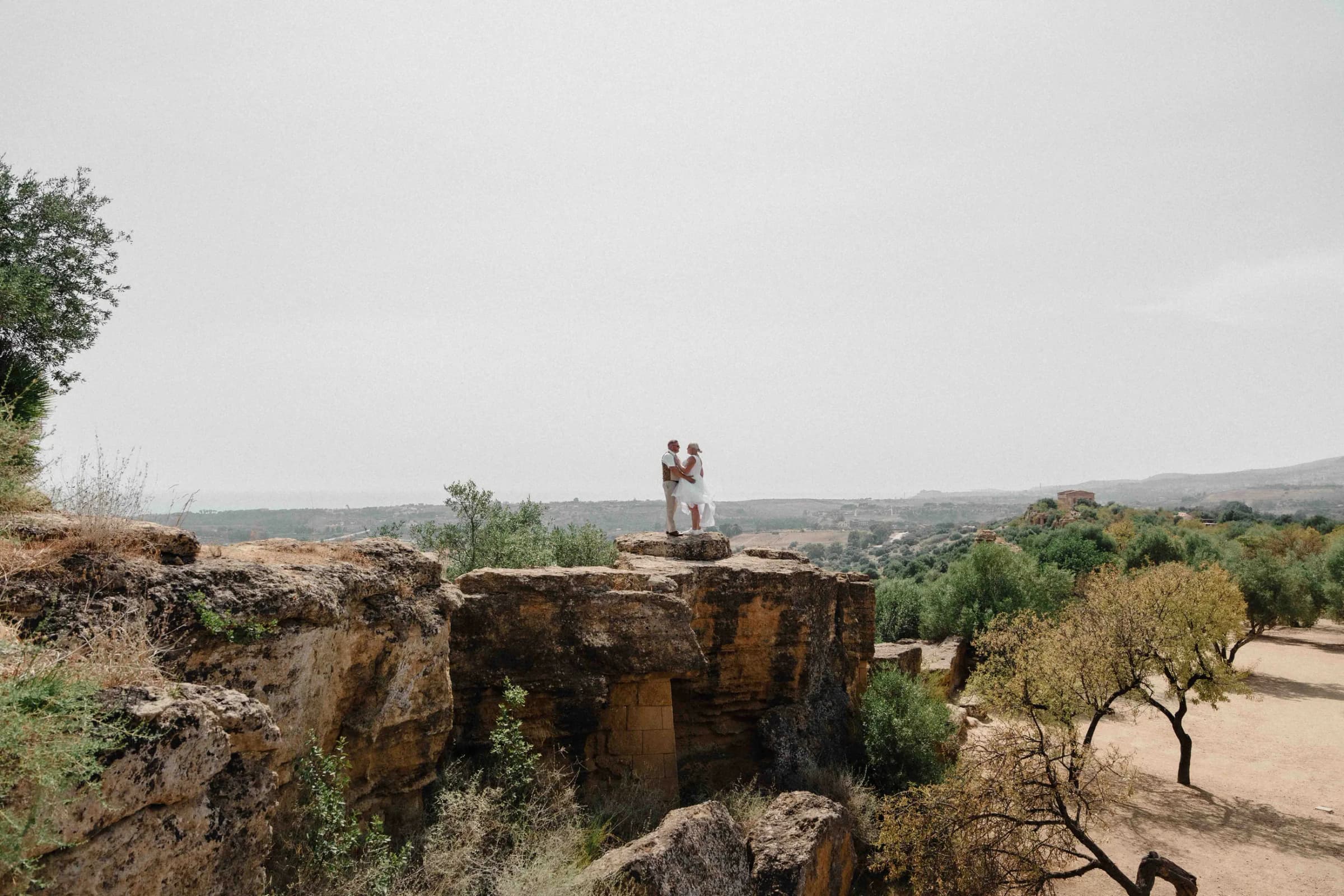 The couple small against a wide valley of olive groves, standing on stone