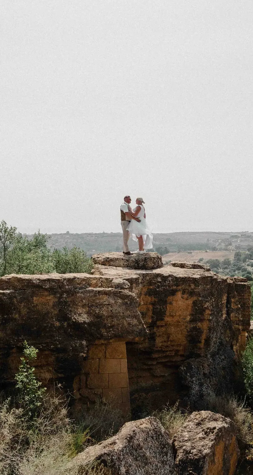 The couple small against a wide valley of olive groves, standing on stone